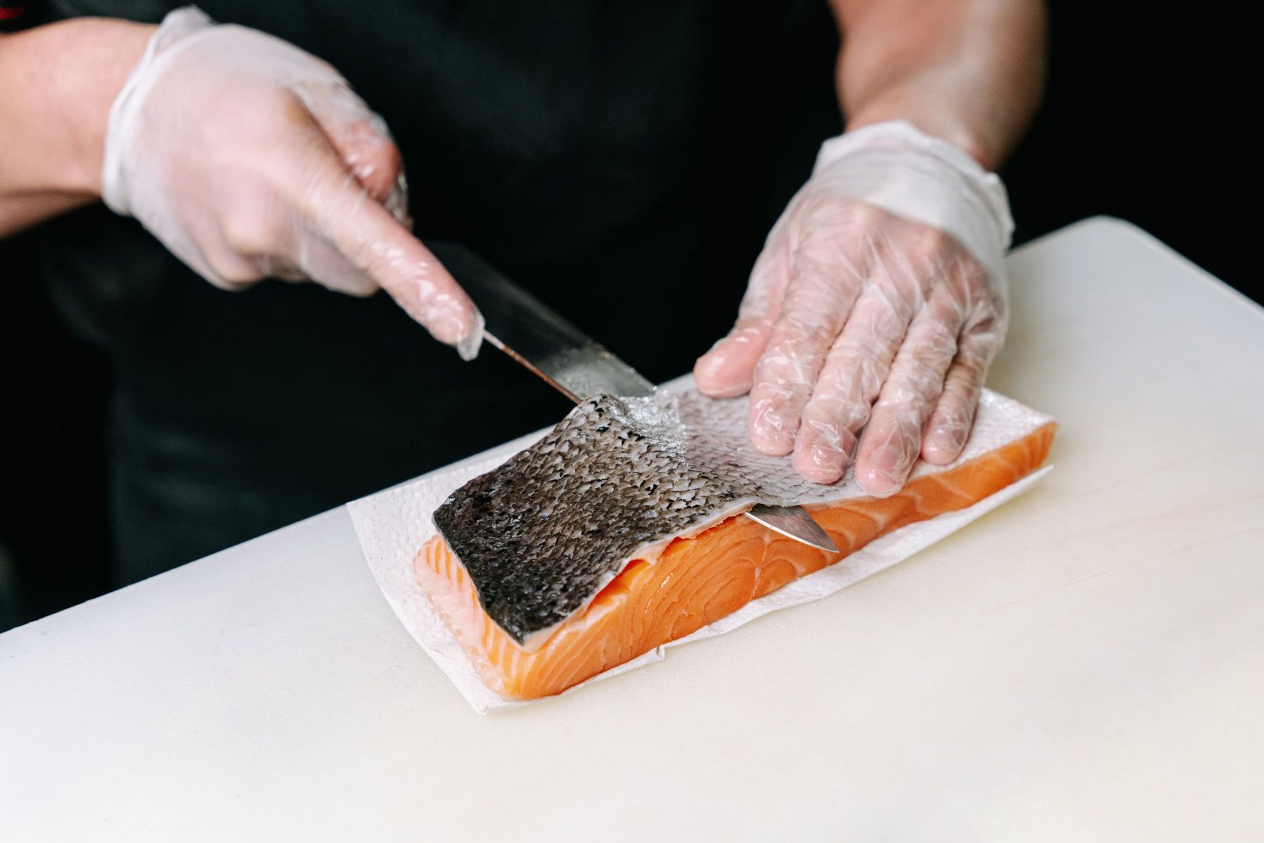 Manos con guantes desechables retirando la piel a un lomo de salmón crudo sobre una tabla blanca