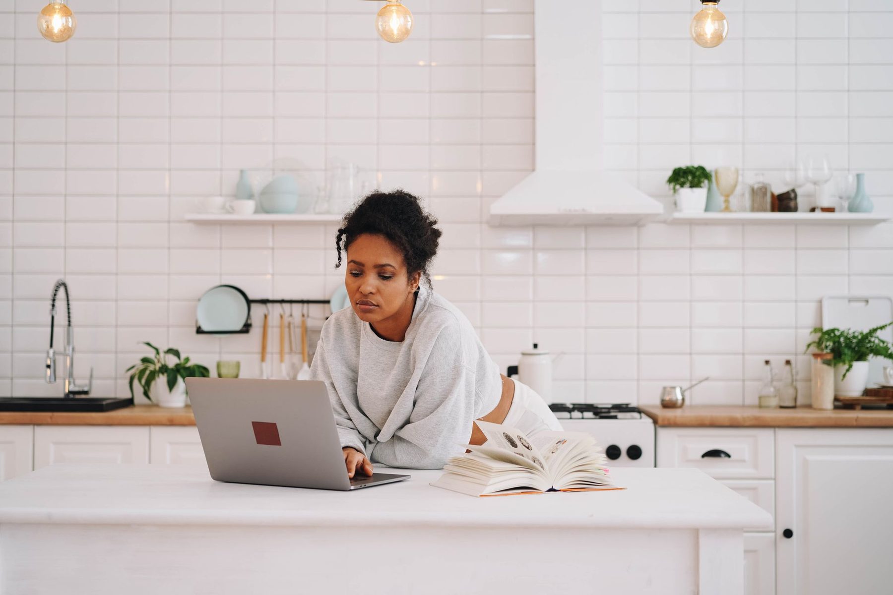Mujer estudiando con un portátil y un libro abierto sobre la encimera de una cocina luminosa