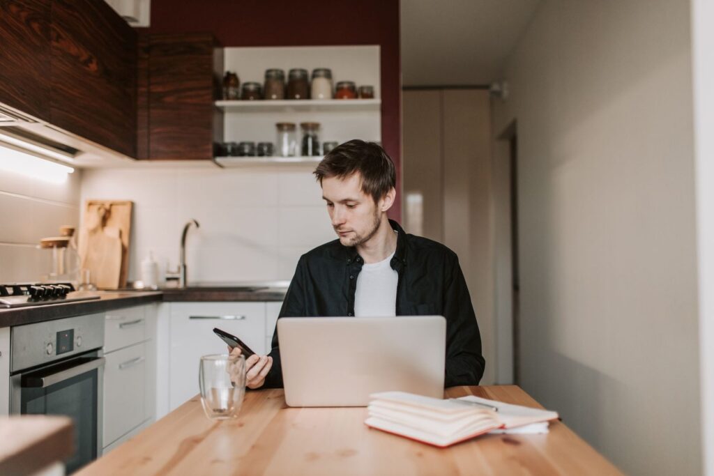 Hombre estudiando en la mesa de su cocina con un portátil, un móvil y un cuaderno abierto