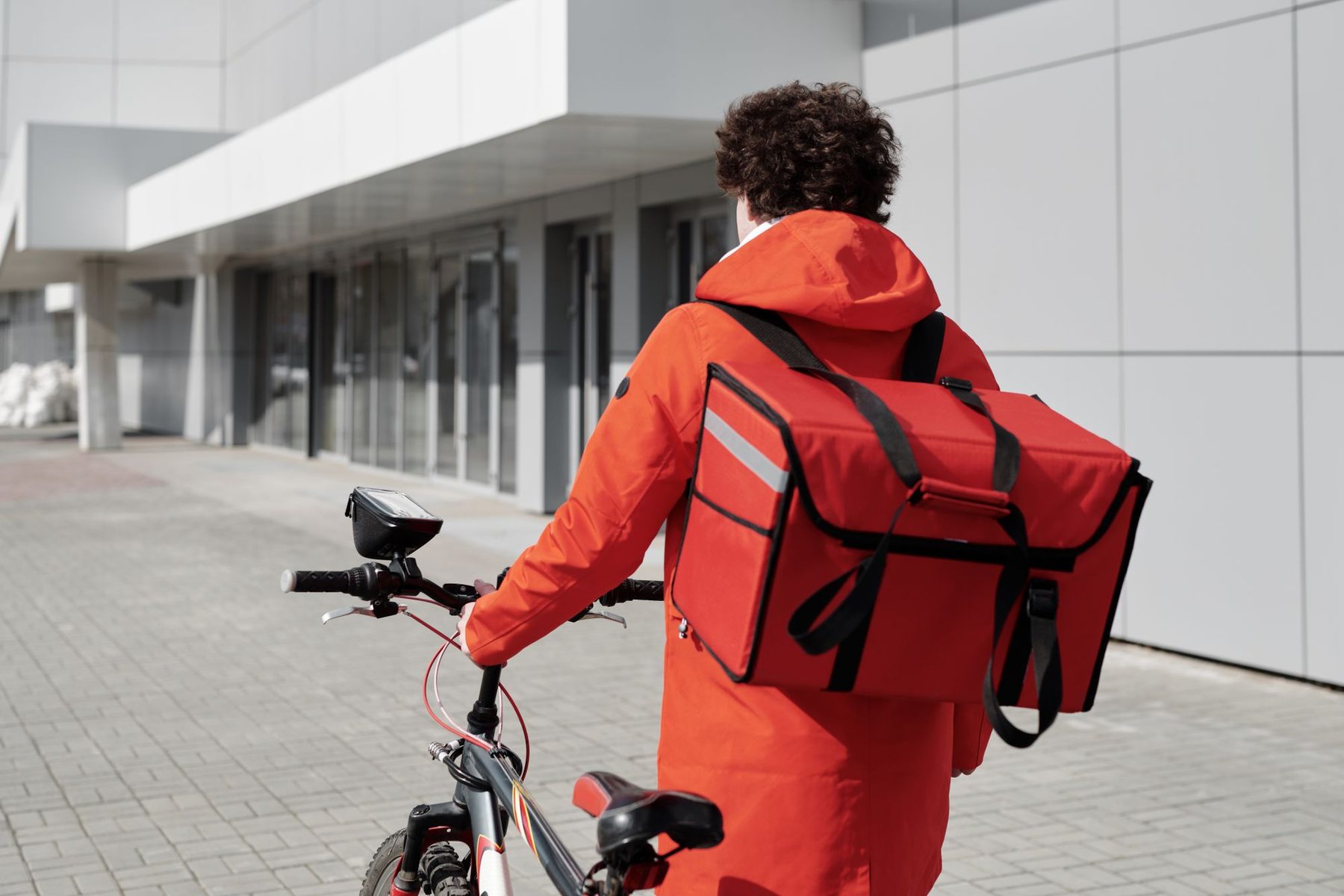 Repartidor de comida de espaldas con una mochila térmica roja caminando junto a su bicicleta