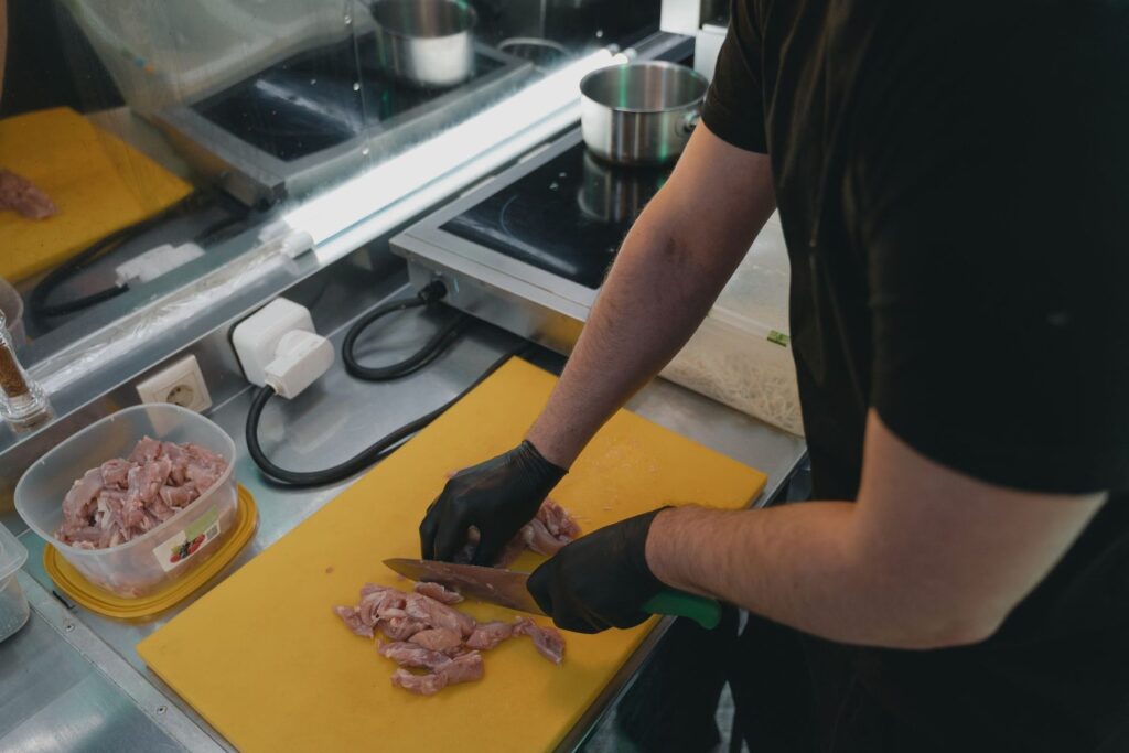 Manipulador con guantes negros cortando carne cruda sobre una tabla de cortar amarilla, con un recipiente aparte