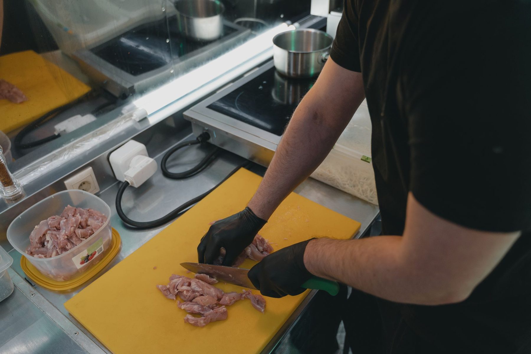 Manipulador con guantes negros cortando carne cruda sobre una tabla de cortar amarilla, con un recipiente aparte